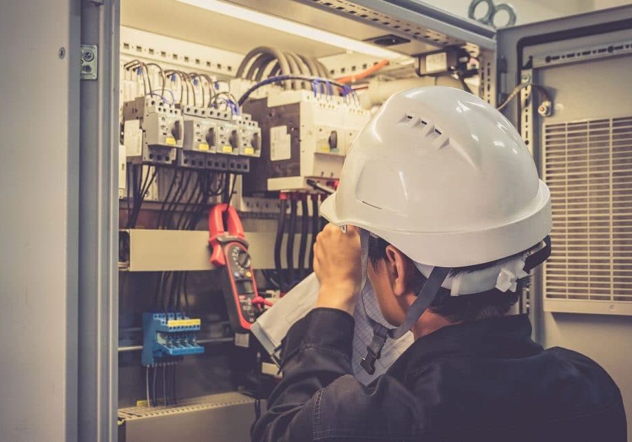 Electrician is measuring voltage or current by voltmeter in control panel of power plant, selective focus on safety helmet